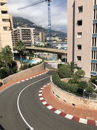 A street scene featuring a sharp corner with red and white curb markings, palm trees, and large residential buildings. A blue construction crane is visible in the background, with a hillside dotted with buildings and cloudy sky behind.