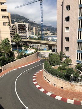 A street scene featuring a sharp corner with red and white curb markings, palm trees, and large residential buildings. A blue construction crane is visible in the background, with a hillside dotted with buildings and cloudy sky behind.