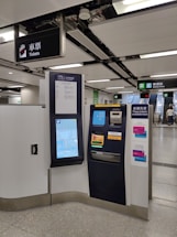 A modern ticket vending machine is situated in a clean subway station. There are clear signage and escalators leading to an exit marked 'Rodney Street'. The machine features a touchscreen interface displaying a map and options for purchasing tickets.