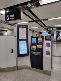 A modern ticket vending machine is situated in a clean subway station. There are clear signage and escalators leading to an exit marked 'Rodney Street'. The machine features a touchscreen interface displaying a map and options for purchasing tickets.
