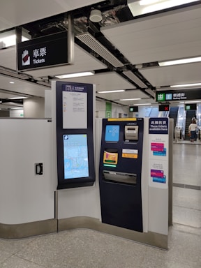 A modern ticket vending machine is situated in a clean subway station. There are clear signage and escalators leading to an exit marked 'Rodney Street'. The machine features a touchscreen interface displaying a map and options for purchasing tickets.