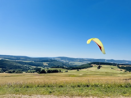A paraglider with a bright yellow and red wing soars over a vast, open landscape. Rolling hills stretch into the distance under a clear blue sky, with patches of green fields and lush forests making up the scenic view.
