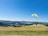 A paraglider with a bright yellow and red wing soars over a vast, open landscape. Rolling hills stretch into the distance under a clear blue sky, with patches of green fields and lush forests making up the scenic view.