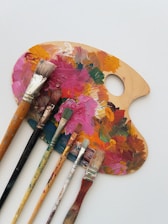 A wooden painter's palette covered in various vibrant acrylic paint colors is placed on a white surface. Several paintbrushes with different sizes and shapes, some stained with paint, are laid across the palette.
