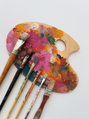 A wooden painter's palette covered in various vibrant acrylic paint colors is placed on a white surface. Several paintbrushes with different sizes and shapes, some stained with paint, are laid across the palette.