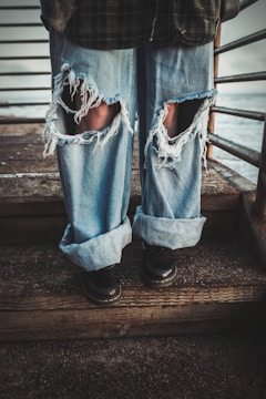A person wearing heavily distressed blue jeans with large rips on the knees, standing on wooden steps next to a metal railing. The jeans are loose and rolled up at the bottom, paired with black shoes.