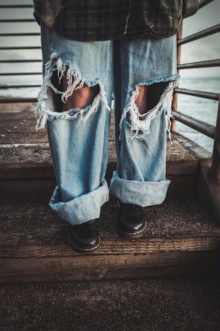 A person wearing heavily distressed blue jeans with large rips on the knees, standing on wooden steps next to a metal railing. The jeans are loose and rolled up at the bottom, paired with black shoes.