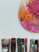 A collection of paintbrushes with varying sizes and colors is arranged in a row below a painter’s palette. The palette displays thick, textured paint in shades of pink, orange, and brown.