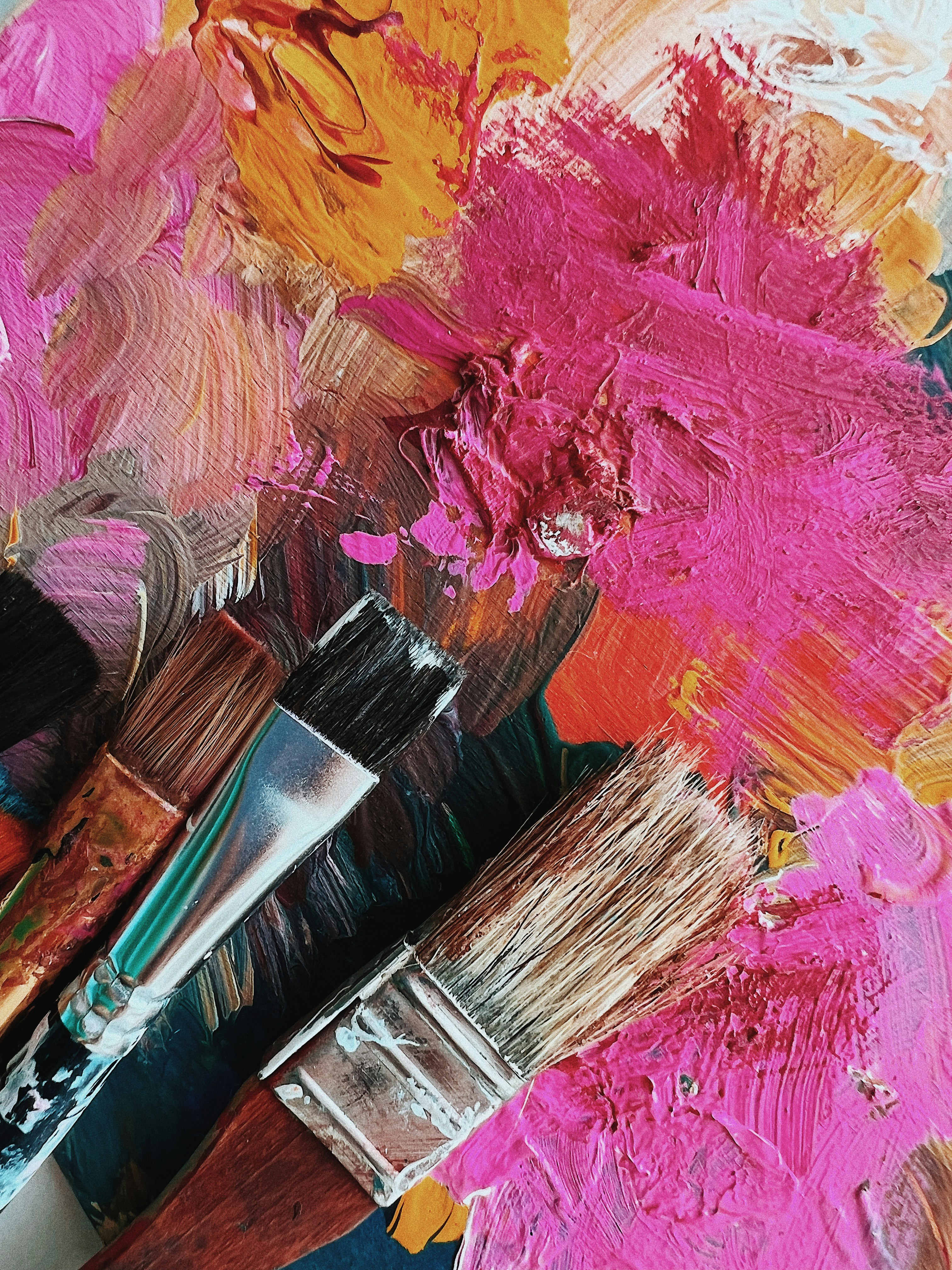 Three paintbrushes with different bristle types rest on a palette covered with thick, textured layers of paint in various colors. The paint is applied in broad, swirling strokes, creating an abstract and vibrant background.