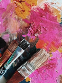 Three paintbrushes with different bristle types rest on a palette covered with thick, textured layers of paint in various colors. The paint is applied in broad, swirling strokes, creating an abstract and vibrant background.