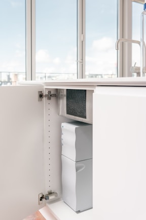An open kitchen cabinet reveals a water filtration or purification system installed inside. The space is well-lit and minimalistic with a modern faucet visible on the counter above.