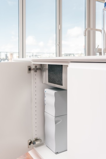 An open kitchen cabinet reveals a water filtration or purification system installed inside. The space is well-lit and minimalistic with a modern faucet visible on the counter above.