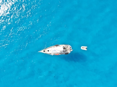 Aerial view of a sailboat anchored in crystal-clear blue water with a small dinghy floating nearby. The sun reflects off the water, creating a serene and peaceful atmosphere.