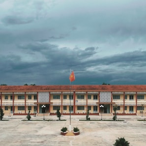 A large, rectangular school building with a red tile roof and yellow walls is central in the image. In front of the building, there is a concrete courtyard with a flagpole featuring a raised red flag with a yellow star. The sky above is covered with dense, dark clouds, suggesting an overcast day. The courtyard is bordered by small trees and a few streetlamps.