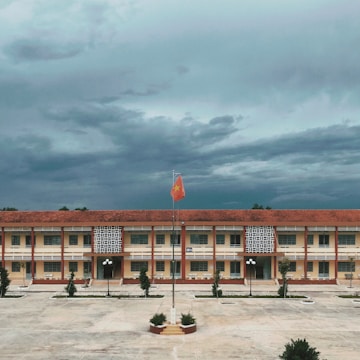 A large, rectangular school building with a red tile roof and yellow walls is central in the image. In front of the building, there is a concrete courtyard with a flagpole featuring a raised red flag with a yellow star. The sky above is covered with dense, dark clouds, suggesting an overcast day. The courtyard is bordered by small trees and a few streetlamps.