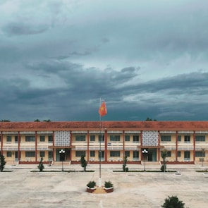 A large, rectangular school building with a red tile roof and yellow walls is central in the image. In front of the building, there is a concrete courtyard with a flagpole featuring a raised red flag with a yellow star. The sky above is covered with dense, dark clouds, suggesting an overcast day. The courtyard is bordered by small trees and a few streetlamps.