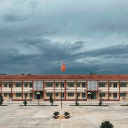A large, rectangular school building with a red tile roof and yellow walls is central in the image. In front of the building, there is a concrete courtyard with a flagpole featuring a raised red flag with a yellow star. The sky above is covered with dense, dark clouds, suggesting an overcast day. The courtyard is bordered by small trees and a few streetlamps.