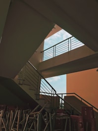 A staircase with metal railings under natural light, showing geometric shapes created by the railings and shadows. An open sky is visible through an angular gap, and stacked furniture or construction materials are placed at the base.