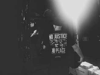 A person stands in a dimly lit room wearing a black t-shirt with white text that reads 'NO JUSTICE NO PEACE.' The image has a high contrast, emphasizing shadows and a bright light source in the background. A box labeled 'GILDAN' is visible on the floor.