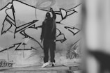 A person in a dark outfit is standing in front of a large wall covered in graffiti art. The artwork consists of bold, abstract shapes with thick black outlines, and the individual appears to be in the process of adding to the graffiti. The setting is urban, with concrete flooring and a sense of movement in the brushstrokes.