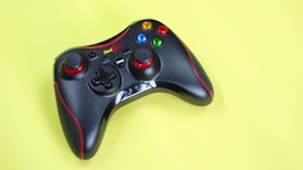 A black game controller with red accents, featuring colorful buttons, is placed against a bright yellow background. The controller has a sleek design with a combination of buttons, triggers, and directional pad.