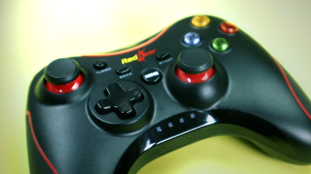 A black gaming controller with red accents rests on a light surface. It features two thumb sticks, a D-pad, and several colorful buttons, including buttons labeled START and BACK, and a logo reading 'Red Star'.