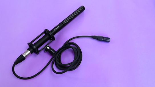 A handheld microphone with a black handle and a coiled black cable connected to it. The microphone and cable are placed on a smooth lavender surface.