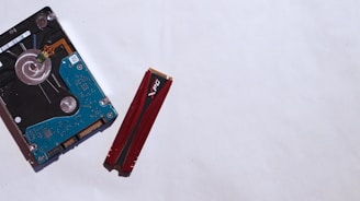 A hard drive and a red RAM stick are placed on a light surface. The hard drive has visible circuitry with a blue circuit board, while the RAM stick features a red casing and is positioned vertically.