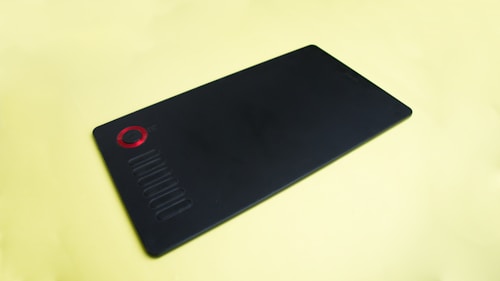 A black graphic tablet with a red dial and several buttons on its surface, resting on a light yellow background.