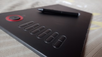 A black graphics tablet with several buttons and a red circular dial on the left side. A stylus pen rests on the surface, providing a sense of readiness for digital creation. The background consists of a soft, beige fabric with subtle patterns.