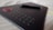 A black graphics tablet with several buttons and a red circular dial on the left side. A stylus pen rests on the surface, providing a sense of readiness for digital creation. The background consists of a soft, beige fabric with subtle patterns.