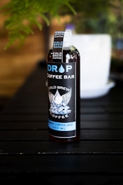 A bottle labeled 'Drop Coffee Bar' stands on a dark wooden surface. The label features the words 'Cold Brew Coffee' with an illustration of wings and ice cubes. The bottle is filled with a dark liquid, likely coffee, and a visible white cap is secured on top. A blurred white object is in the background amidst green foliage.