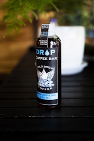 A bottle labeled 'Drop Coffee Bar' stands on a dark wooden surface. The label features the words 'Cold Brew Coffee' with an illustration of wings and ice cubes. The bottle is filled with a dark liquid, likely coffee, and a visible white cap is secured on top. A blurred white object is in the background amidst green foliage.