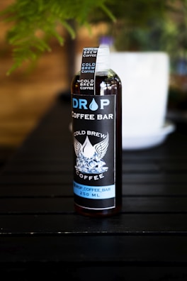 A bottle labeled 'Drop Coffee Bar' stands on a dark wooden surface. The label features the words 'Cold Brew Coffee' with an illustration of wings and ice cubes. The bottle is filled with a dark liquid, likely coffee, and a visible white cap is secured on top. A blurred white object is in the background amidst green foliage.