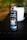 A bottle labeled 'Drop Coffee Bar' stands on a dark wooden surface. The label features the words 'Cold Brew Coffee' with an illustration of wings and ice cubes. The bottle is filled with a dark liquid, likely coffee, and a visible white cap is secured on top. A blurred white object is in the background amidst green foliage.