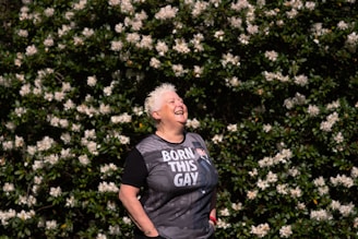 A person with short white hair is laughing joyfully while standing in front of a lush green bush covered with numerous white flowers. The person is wearing a black T-shirt with text on it and appears to be enjoying the moment.
