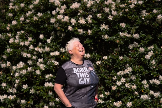 A person with short white hair is laughing joyfully while standing in front of a lush green bush covered with numerous white flowers. The person is wearing a black T-shirt with text on it and appears to be enjoying the moment.