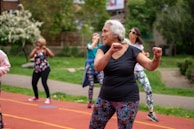 A group of people is participating in an outdoor exercise class on a track. The focus is on an older woman with gray hair wearing a black shirt and colorful leggings, enthusiastically performing workout movements. In the background, several other women are similarly dressed in activewear, exercising with determination. The setting appears to be in a park with lush green trees and bushes.