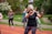 A group of people is participating in an outdoor exercise class on a track. The focus is on an older woman with gray hair wearing a black shirt and colorful leggings, enthusiastically performing workout movements. In the background, several other women are similarly dressed in activewear, exercising with determination. The setting appears to be in a park with lush green trees and bushes.
