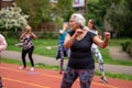 A group of people is participating in an outdoor exercise class on a track. The focus is on an older woman with gray hair wearing a black shirt and colorful leggings, enthusiastically performing workout movements. In the background, several other women are similarly dressed in activewear, exercising with determination. The setting appears to be in a park with lush green trees and bushes.