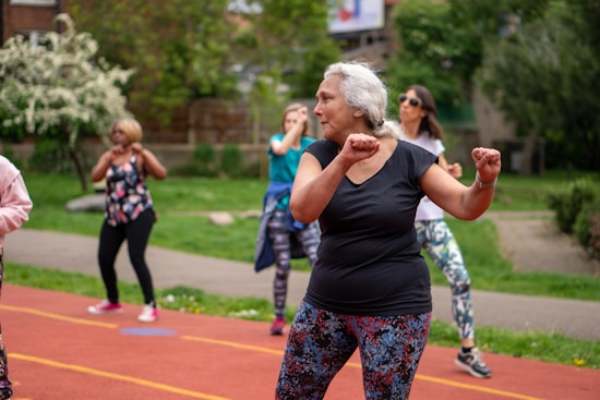 A group of people is participating in an outdoor exercise class on a track. The focus is on an older woman with gray hair wearing a black shirt and colorful leggings, enthusiastically performing workout movements. In the background, several other women are similarly dressed in activewear, exercising with determination. The setting appears to be in a park with lush green trees and bushes.