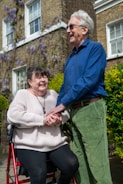 An elderly couple is smiling and holding hands outdoors. One is seated on a mobility aid, wearing a beige sweater and dark pants. The other stands wearing a blue shirt and green trousers. They are in front of a brick building adorned with flowering vines and surrounded by lush greenery.