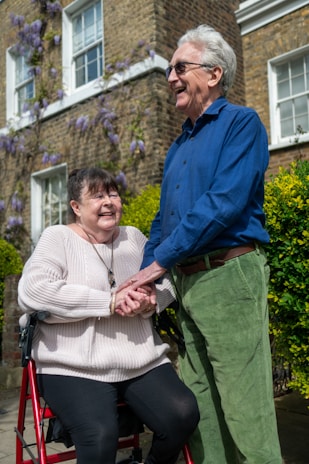 An elderly couple is smiling and holding hands outdoors. One is seated on a mobility aid, wearing a beige sweater and dark pants. The other stands wearing a blue shirt and green trousers. They are in front of a brick building adorned with flowering vines and surrounded by lush greenery.