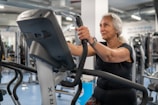 An elderly person is exercising on a stationary bike in a gym setting. The environment is filled with various gym equipment and modern fitness machines. The person appears focused and determined, wearing casual workout attire.