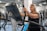 An elderly person is exercising on a stationary bike in a gym setting. The environment is filled with various gym equipment and modern fitness machines. The person appears focused and determined, wearing casual workout attire.