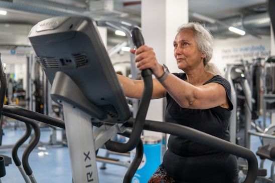 An elderly person is exercising on a stationary bike in a gym setting. The environment is filled with various gym equipment and modern fitness machines. The person appears focused and determined, wearing casual workout attire.