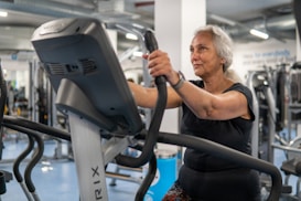 An elderly person is exercising on a stationary bike in a gym setting. The environment is filled with various gym equipment and modern fitness machines. The person appears focused and determined, wearing casual workout attire.