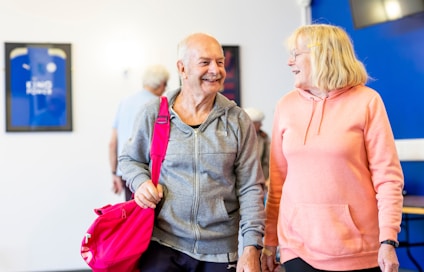 An older man and woman are smiling at each other in a bright room. The man is holding a pink gym bag and wearing a grey jacket, while the woman is in a peach hoodie. In the background, a blurred figure and framed sports jerseys can be seen on the wall.