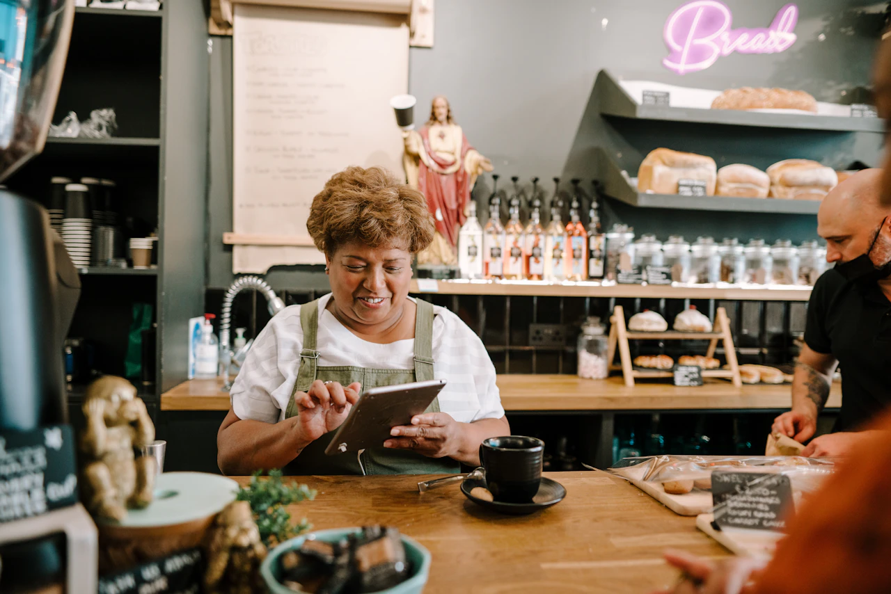 Woman in a green apron smiles at a tablet behind a café counter while a colleague works nearby.