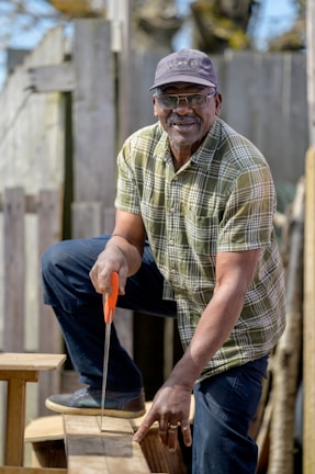 A man wearing a plaid shirt and a cap is using a hand saw to cut a piece of wood. He is outdoors, positioned in front of a wooden fence, and appears to be focused on his task.